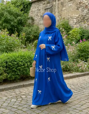 Muslima mit blauem Hijab trägt königsblaue Abaya mit weißen Schleifen von Noor Shop und läuft über einen Kopfsteinweg im Garten