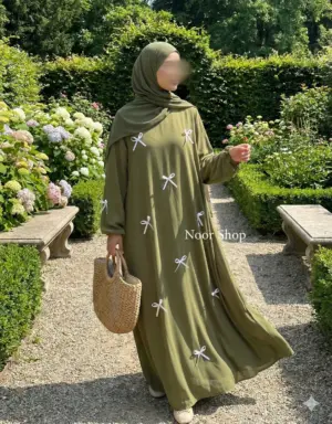 Muslima mit olivgrüner Abaya mit weißen Schleifen von Noor Shop steht in einem Blumengarten und trägt eine Strohtasche
