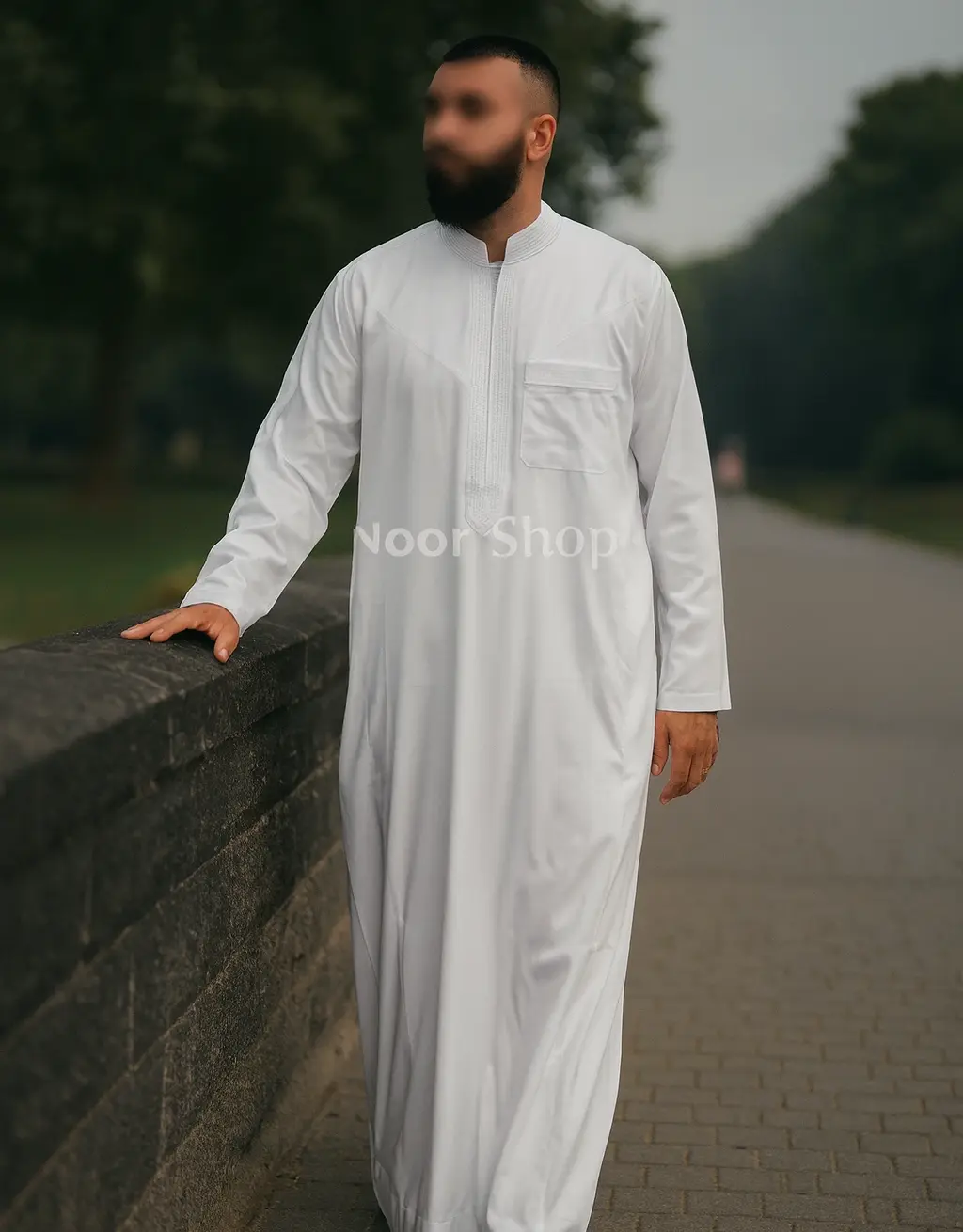 Mann trägt weiße Herren Djellaba mit Stickerei von Noor Shop und läuft an einer Steinmauer im Park