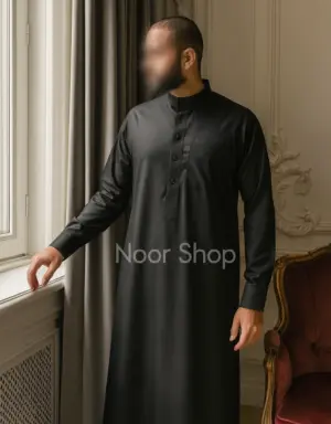 Mann trägt schwarzen Herren Thobe mit Stehkragen und Knopfleiste von Noor Shop und steht am Fenster eines eleganten Raumes