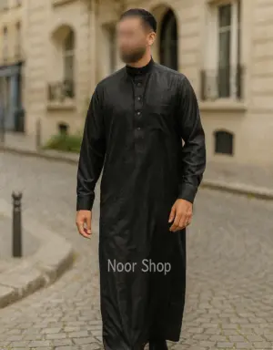 Mann trägt schwarzen Herren Thobe mit Stehkragen, Knopfleiste und Brusttasche von Noor Shop auf einer Kopfsteinpflaster Straße