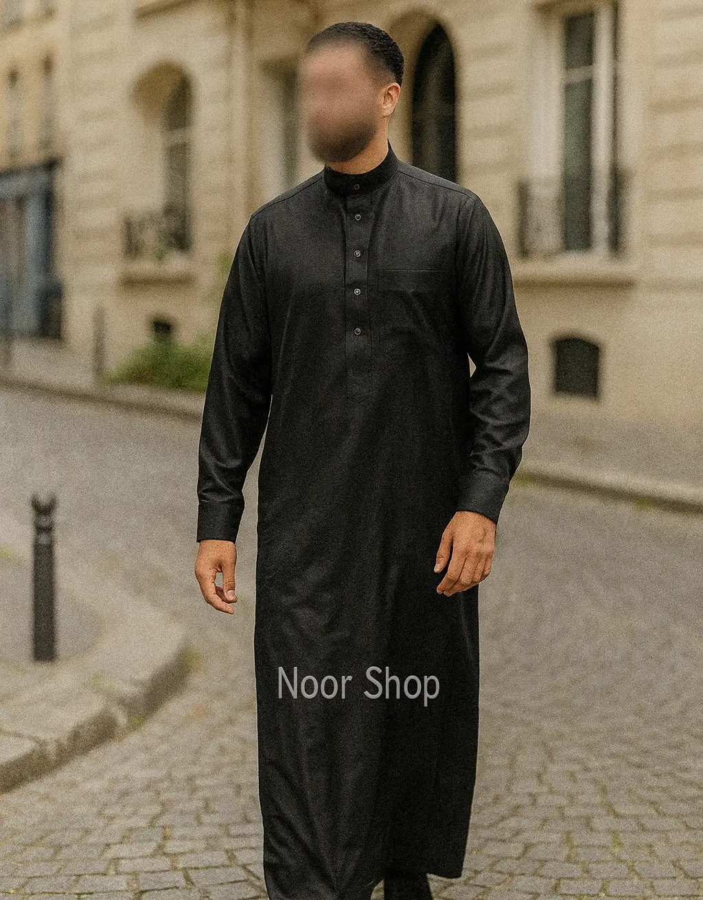 Mann trägt schwarzen Herren Thobe mit Stehkragen, Knopfleiste und Brusttasche von Noor Shop auf einer Kopfsteinpflaster Straße